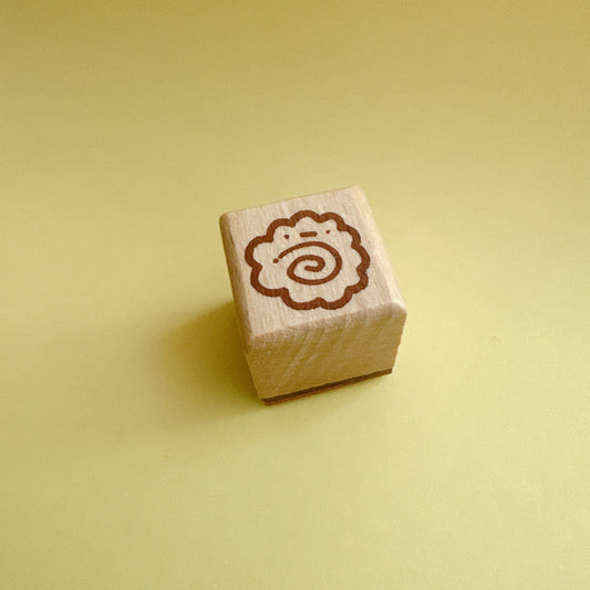A small square wooden stamp block with a brown spiral design on top, photographed on a smooth pale yellow background.