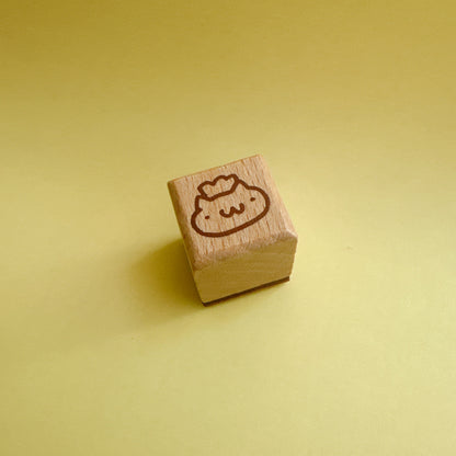 A small wooden cube stamp with a cute smiling cat engraved on top, set against a solid pale yellow background.