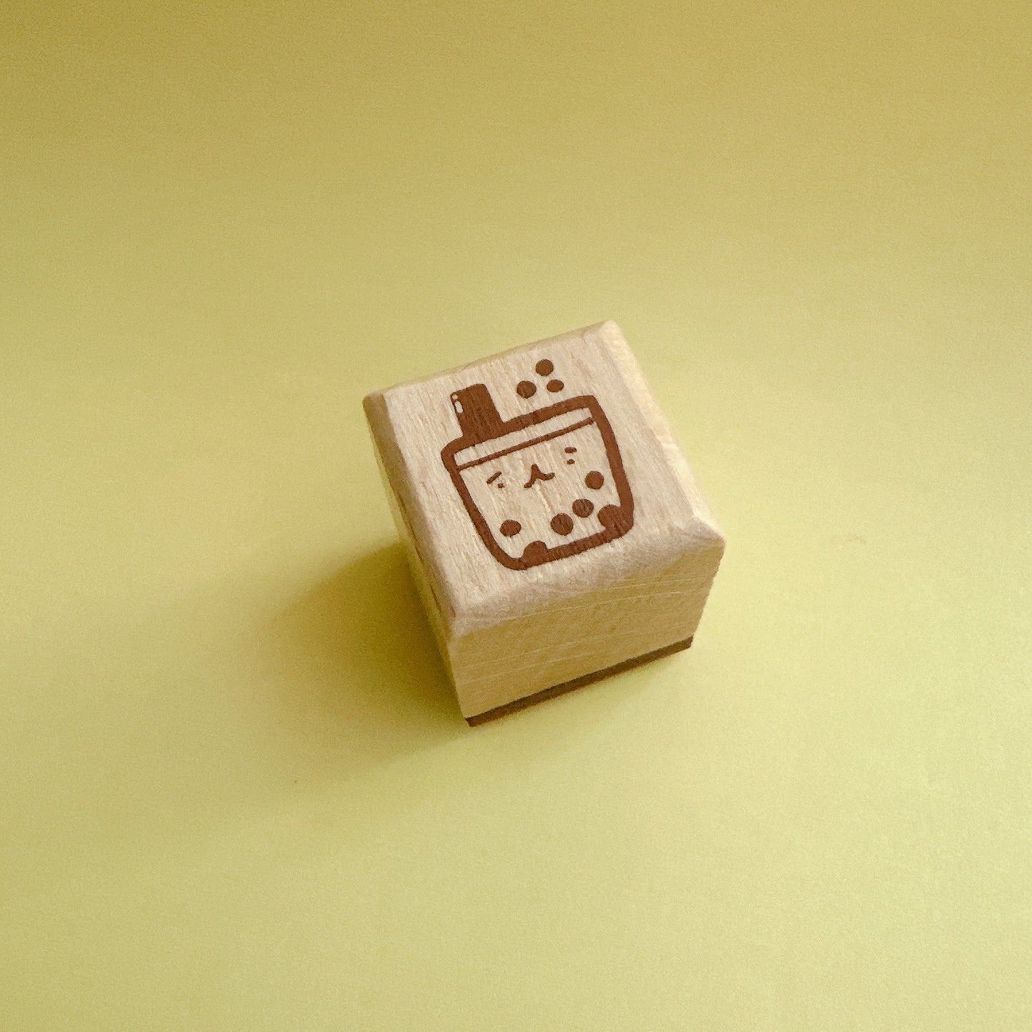 A small wooden rubber stamp with a brown boba tea cup design, standing upright on a solid pale yellow background.