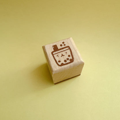 A small wooden rubber stamp with a brown boba tea cup design, standing upright on a solid pale yellow background.