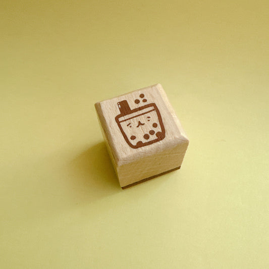 A small wooden rubber stamp with a brown boba tea cup design, standing upright on a solid pale yellow background.