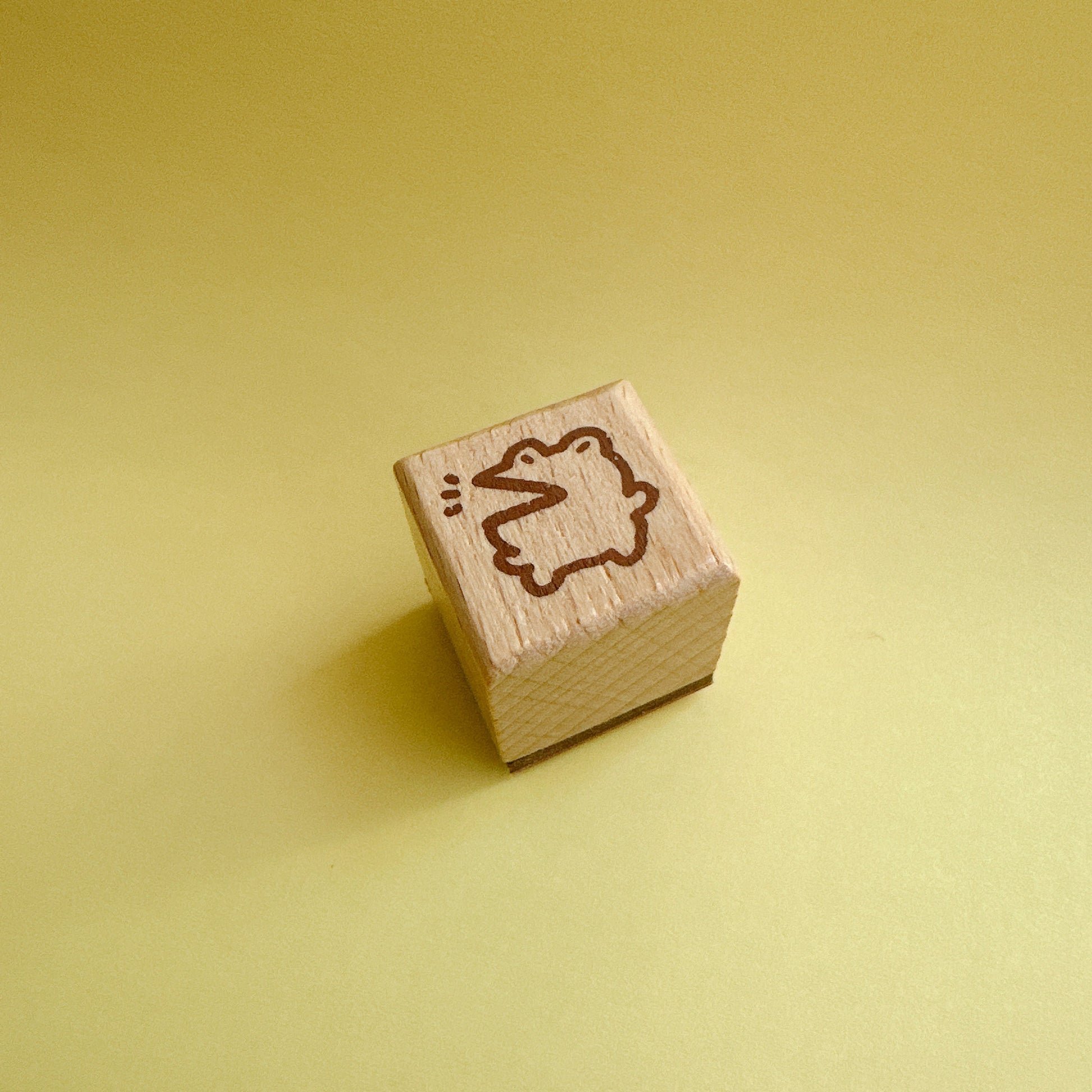 A small wooden cube stamp with a brown engraved outline of a duck, set against a solid pale yellow background.