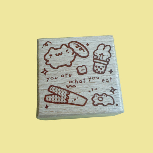 A square wooden block featuring cute food-themed illustrations and the text 'you are what you eat' in brown, shown against a solid pale yellow background.