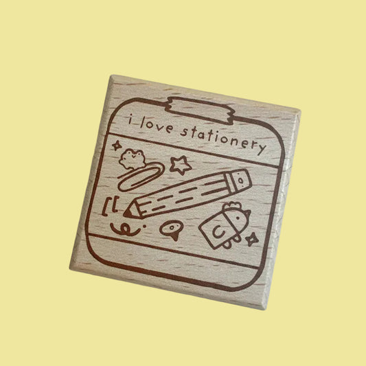 A square wooden rubber stamp with a brown engraved design of stationery items and the text 'I love stationery', shown on a pale yellow-green background.