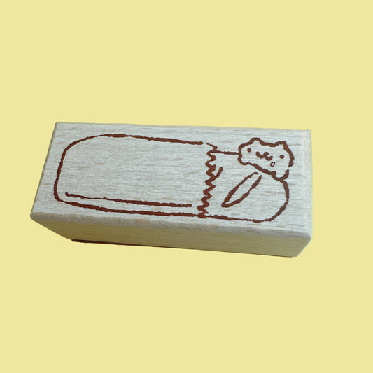 A rectangular wooden stamp block with a simple brown line drawing of a sleeping figure, shown on a pale yellow background.