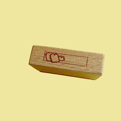 A small rectangular wooden stamp block with a red engraved dotted rectangle and simple figure design, standing upright on a solid yellow background.