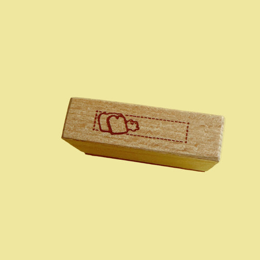 A small rectangular wooden stamp block with a red engraved dotted rectangle and simple figure design, standing upright on a solid yellow background.